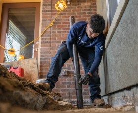 A worker installs a pipe at a construction site with exposed ground, wearing gloves and dark clothing.