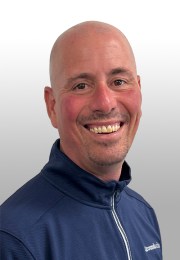 Smiling bald man in a navy zip-up jacket against a light gradient background.