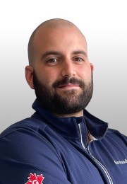 Man with a beard in a navy jacket smiles slightly, standing against a light gray background.