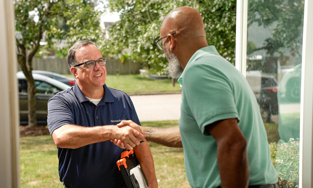 Two men smiling and shaking hands at an open door, one holding a toolbox. Trees and a street are visible outside.