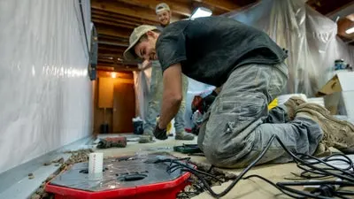 Two men work on a sump pump while kneeling on the floor of a basement, with tools scattered around them.