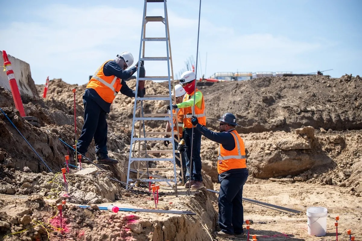 Four construction workers in safety gear position a ladder at a dirt excavation site on a sunny day.