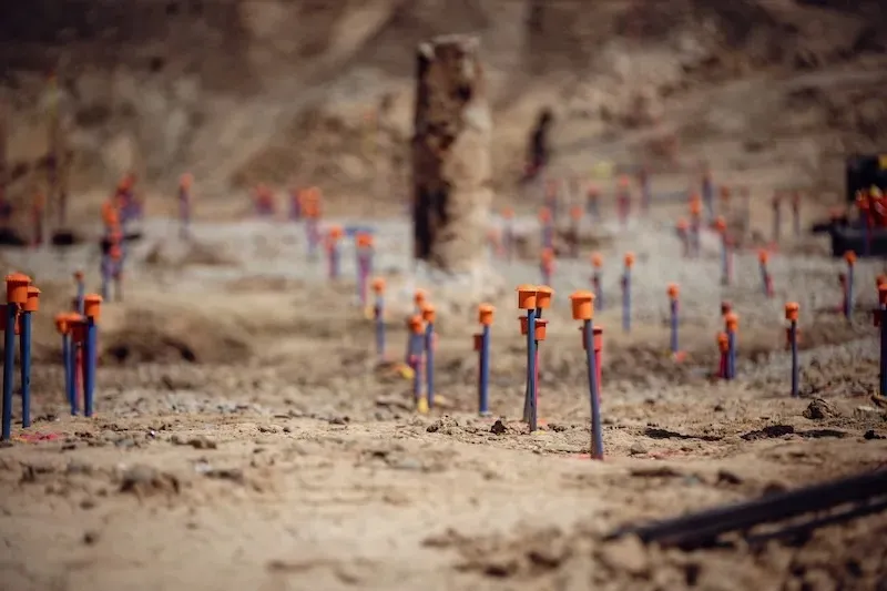 Numerous survey stakes with orange caps are placed in dry, rocky soil at a construction site.