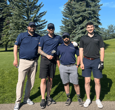 Four men stand together on a golf course in sunny weather, wearing golf attire and smiling at the camera.