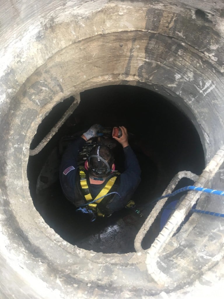 Worker wearing safety gear descends into a large, circular concrete shaft with a rope and ladder.