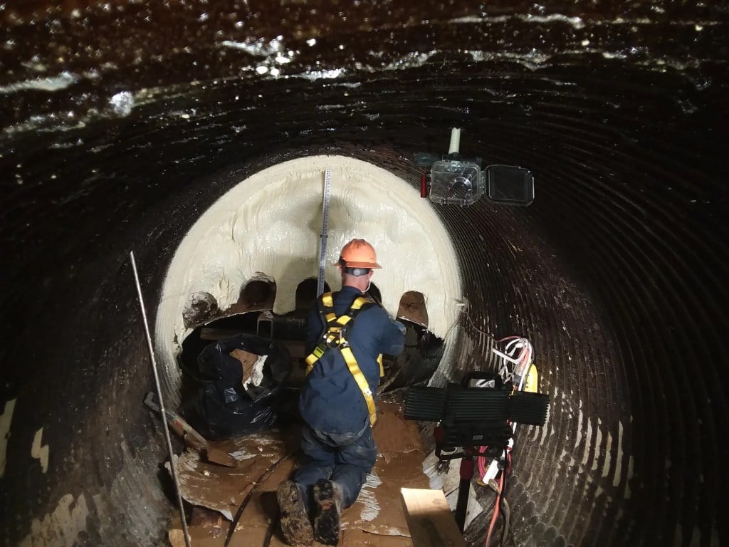 Worker in safety gear applies coating inside a large, dark tunnel using specialized equipment.