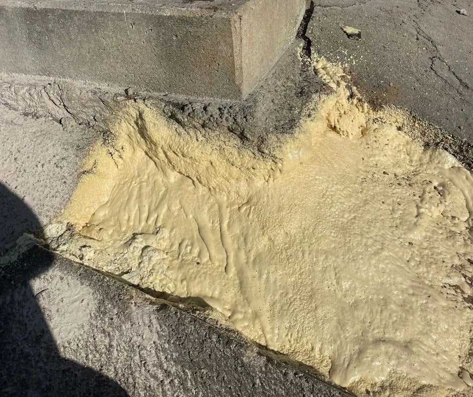 Yellowish foam oozes and hardens on cracked pavement beside a concrete step in bright sunlight.