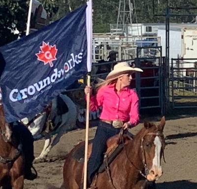 Woman in pink shirt rides horse, holding "Groundworks" flag at an outdoor rodeo event.