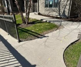 Curved sidewalk and metal fence beside a grassy area in front of a house with trees and shrubs.
