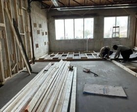 Two people working on wooden framing inside a room under construction with large windows in the background.