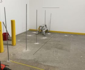 Six metal rods are installed upright in a concrete floor near a step ladder, with yellow bollards at the side.