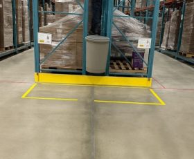 A person stands on a trash can between warehouse shelves with pallets and boxes in the background.
