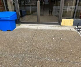 A glass door with a reflection, blue bin on left, scattered debris, and a wooden plank on the right side.